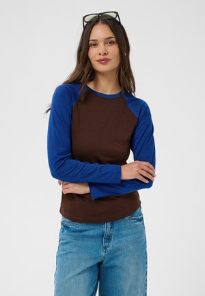 Femme aux longs cheveux foncés, portant une chemise marron avec des manches longues bleues et un jean bleu clair, bras croisés, lunettes de soleil sur la tête.