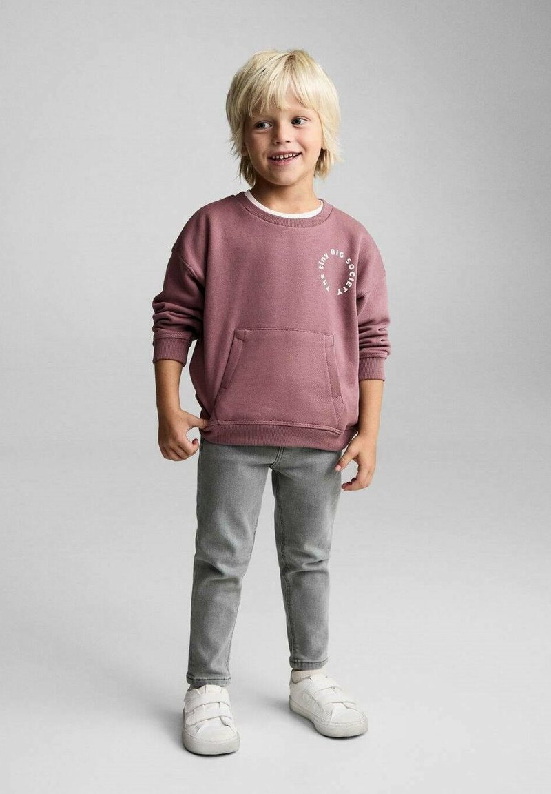 Sweatshirt roz prăfuit cu un grafic alb, buzunar mare în față, combinat cu pantaloni gri pe corp și pantofi slip-on albi.