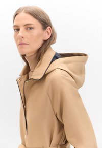 Khaki kapuce s kontrastní tmavou podšívkou, zlatými detaily a měkkou strukturou. Detailní záběr modelu, který ji nosí na světlém pozadí.