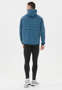 Chaqueta deportiva azul con parte superior trasera acolchada, capucha ajustable y acentos reflectantes, combinada con leggins negros ceñidos y zapatillas de running.