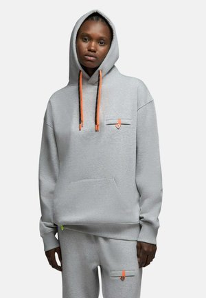 Personne noire portant un sweat à capuche gris clair et un pantalon assorti avec des cordons orange et noir et de petites poches boutonnées, se tenant devant un fond blanc.