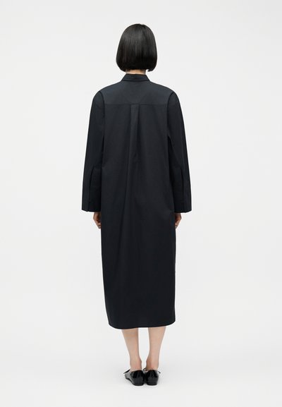 Γυναίκα με κοντά μαύρα μαλλιά φοράει μακρύ μαύρο φόρεμα με κουμπιά και μαύρα επίπεδα παπούτσια, στέκεται με την πλάτη προς το λευκό φόντο.