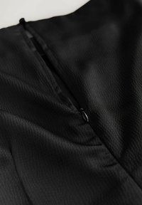 Fekete ruhaanyag textúrált felülettel, hátul cipzáras záródással és diszkrét redőzött részletekkel a nyakvonalnál.