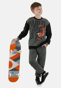 Jongen in een grijze en zwarte sweatshirt met tijgerdesign, grijze broek en zwarte sneakers, die een oranje en grijze skateboard vasthoudt.