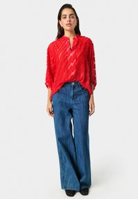 Top rosso, trasparente, con texture, scollatura a V e maniche a tre quarti abbinato a jeans in denim blu a vita alta e gamba larga. Scarpe nere visibili.