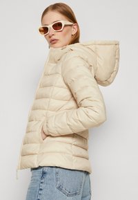 Beige polstret jakke med hette, med kviltet tekstur og elastiske mansjetter. Kombinert med lyseblå jeans og hvite solbriller.