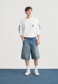 Λευκό πουκάμισο με μακριά μανίκια και μπλε γραφικό στο μανίκι, συνδυασμένο με μπλε τζιν σορτς και λευκά αθλητικά παπούτσια με πολύχρωμες λεπτομέρειες. Απλό φόντο.