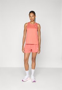 Conjunto de camiseta sin mangas y pantalones cortos en color coral, con tela transpirable, detalles de malla y un logotipo en el pecho. Combinado con calcetines blancos y zapatillas deportivas.