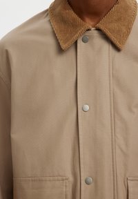 Veste beige avec col en velours côtelé marron, boutons-pression à l'avant et poches supérieures visibles, portée par une personne.