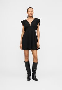 Vrouw draagt een kort zwart jurkje met gerimpelde schouders en een diepe V-hals, gecombineerd met kniehoge zwarte leren laarzen, staand op een witte achtergrond.