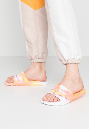 Pieds portant un pantalon de survêtement blanc, beige et orange avec des sandales slides tie-dye jaune, rose et blanche sur un sol clair uni.