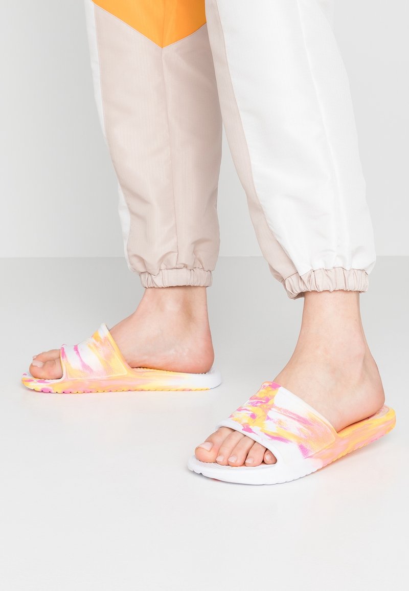 Pieds portant un pantalon de survêtement blanc, beige et orange avec des sandales slides tie-dye jaune, rose et blanche sur un sol clair uni.