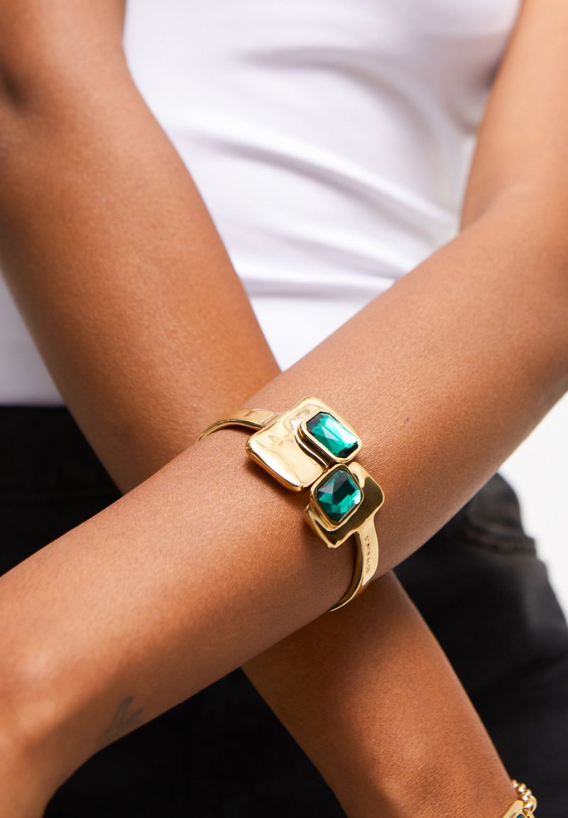 Brazalete de oro con dos piedras facetadas de color verde esmeralda, superficie lisa, diseño abierto con extremos curvados, llevado en un brazo desnudo.