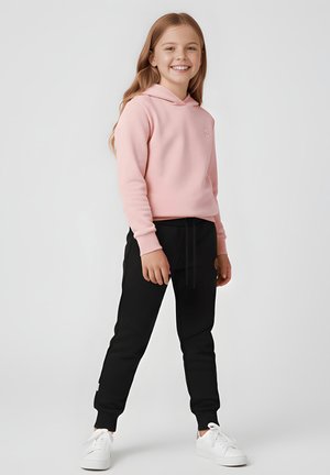 Uśmiechnięta dziewczyna o długich włosach w jasnoróżowej bluzie z kapturem, czarnych spodniach dresowych i białych trampkach stoi na tle jednolitego tła.