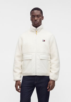 Junger Mann trägt eine weiße Fleecejacke mit Reißverschluss und großen Fronttaschen sowie dunkle Hose, steht vor einfachem Hintergrund.
