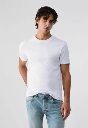 Giovane uomo con capelli scuri e ricci che indossa una maglietta bianca semplice a maniche corte e jeans azzurri su uno sfondo grigio uniforme.