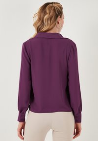 Blusa morada de manga larga con cuello, puños abotonados y una textura suave. Presenta un ligero drapeado y un corte ajustado.