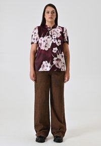 Top viola a maniche corte con scollatura a V abbinato a pantaloni larghi marroni e scarpe chunky nere su sfondo neutro.