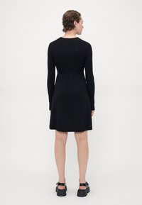 Svart ribbestrikket kjole med lange ermer og tettsittende overdel, som vidder seg ut ved midjen. Kjolens kant sitter over kneet. Brukt med kraftige svarte sandaler.