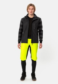 Mannen buitenoutfit: zwarte jas met grijs geruite mouwen, neongele shorts en zwarte leggings. Kenmerken zijn ritszakken en contrasterende kleuren.
