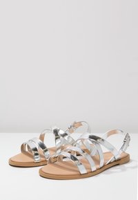 Sandali metallici argento con più fasce larghe e suola in gomma color cammello. Presentano una cinghietta alla caviglia regolabile con fibbia.