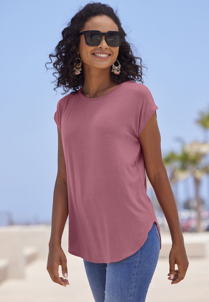 Pink short-sleeve t-shirt with a round neckline, side slits, and a relaxed fit, paired with blue jeans and large black sunglasses.