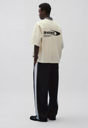 Mladý muž stojaci zboku, oblečený v krémovej košeli s textom „SHAPES“ na chrbte a široké čierne nohavice so širokými bielymi bočnými pruhmi.