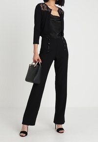 Černý krátký kardigan s krajkovými detaily, saténový top, černé kalhoty s vysokým pasem a knoflíkovými ozdobami, doplněné malou černou kabelkou.