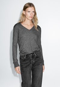 Maglietta grigia a maniche lunghe con scollo a V realizzata in tessuto morbido, abbinata a jeans neri a vita alta. Il design presenta una vestibilità rilassata e polsini a costine.