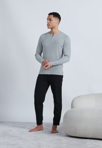 Άνδρας που στέκεται ξυπόλητος σε δάπεδο με χαλί, φοράει γκρι μπλούζα με μακριά μανίκια και μαύρο παντελόνι δίπλα σε μια εκρού καμπυλωτή καναπέ.