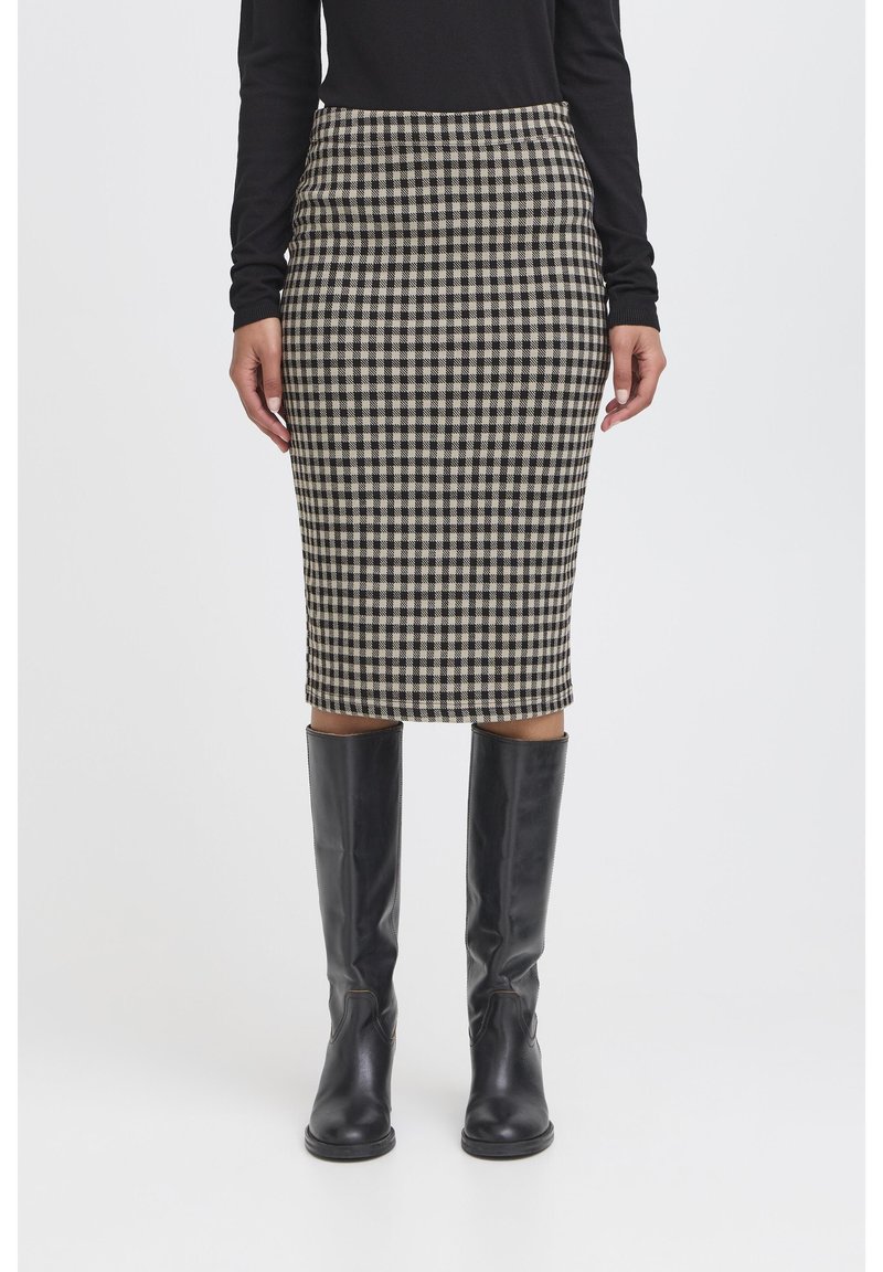 Frau trägt einen schwarz-weiß karierten Bleistiftrock, ein schwarzes langärmliges Oberteil und kniehohe schwarze Lederstiefel, steht vor weißem Hintergrund.