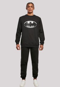 Felpa nera con cappuccio e pantaloni della tuta abbinati, decorati con un grande logo di Batman in bianco. Presenta un colletto rotondo e polsini a costine. Indossati con sneakers bianche.