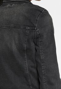 Persona che indossa una giacca di denim nera sbiadita, vista dalla parte superiore della schiena e dalla spalla, su uno sfondo chiaro uniforme.