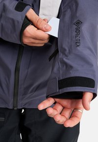 Persoon in een paarse Gore-Tex jas steekt een witte kaart in een met rits afgesloten mouwzakje, waarbij de mouwdetails met klittenbandsluiting zichtbaar zijn.