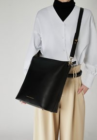 Svart läderaxelväska med en platt, fyrkantig form; har guldfärgade metallaccenter och en präglad logotyp längst ner.