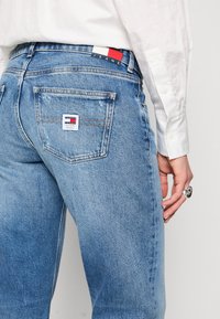 Svetle modré džíny z denimu s vybledlým vzhledem, obsahující obdélníkovou nášivku s vícebarevným logem a zadní kapsy.