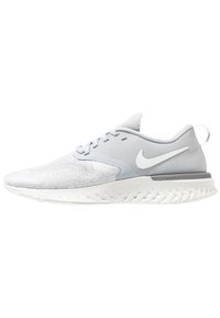 Zapatilla deportiva de color gris claro con parte superior texturizada, acentos lisos y suela blanca con un patrón ondulado. Logotipo de Nike en el costado.