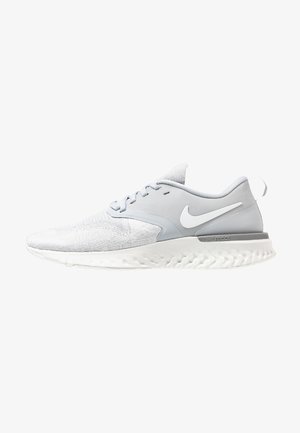 Scarpa sportiva grigia chiara con una parte superiore testurizzata, dettagli lisci e una suola bianca con un motivo ondulato. Logo Nike sul lato.