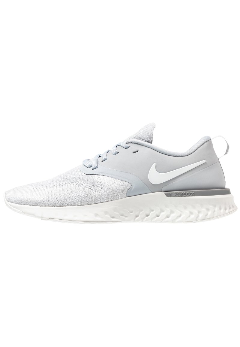 Zapatilla deportiva de color gris claro con parte superior texturizada, acentos lisos y suela blanca con un patrón ondulado. Logotipo de Nike en el costado.
