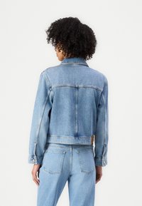 Giacca di jeans azzurro chiaro con design corto, chiusura a bottoni e due tasche posteriori. Presenta un colletto e dettagli di cucitura visibili.