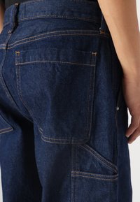Pantalones de mezclilla azul oscuro con costuras naranjas, que cuentan con bolsillos traseros y un bolsillo lateral de utilidad. La tela tiene una textura resistente.