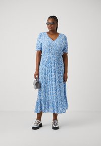 Μπλε φλοράλ μέγιστο φόρεμα με φουσκωτά μανίκια, V-λαιμόκοψη, σμιλευμένη μέση και κρεμαστό φούστα. Συνδυάζεται με λαμπερές ασημένιες πλατφόρμες και γκρι τσάντα.