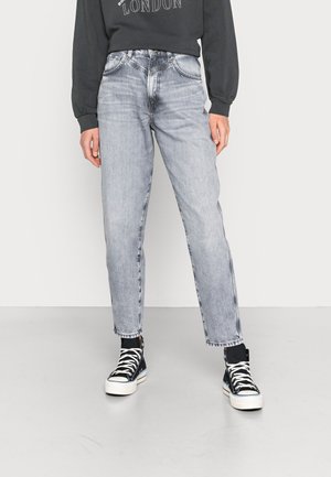 Osoba ubrana w wyblakłe szare jeansy o prostych nogawkach, czarną bluzę z napisem "London", czarne wysokie trampki, stojąca na jednolitym tle.
