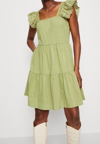 Lichtgroene jurk met een vierkante halslijn, gelaagd ontwerp en mouwen met ruches en kantafwerking. Model poseert met handen in de zakken, draagt crèmekleurige enkellaarzen.