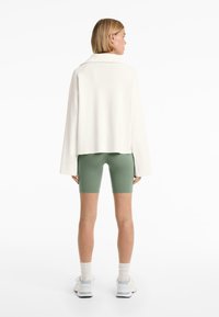Pullover oversized bianco in maglia con un colletto ampio, abbinato a pantaloni bike verdi. Scarpe da ginnastica bianche completano l'outfit. Sfondo semplice.