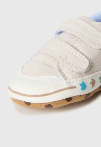 Μπεζ υφασμάτινο παπ ούτσι με δύο ιμάντες βέλκρο, λευκή λαστιχένια σόλα και διακοσμητικά μπλε και καφέ μοτίβα κατά μήκος της άκρης. Υφή με ανάγλυφο σχέδιο.