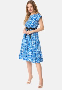 Blauwe bloemenjurk met korte mouwen en een gelaagde rok. Beschikt over een zwarte tailleband en complexe witte patronen op de stof.