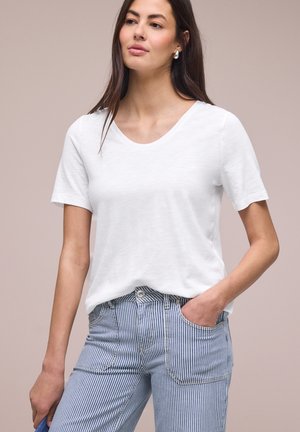 Femme portant un t-shirt blanc à manches courtes et un pantalon à rayures verticales bleu et blanc, avec une main dans la poche, sur un fond uni.