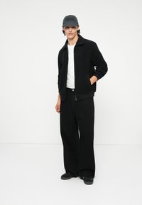 Giacca in pile nera con colletto, t-shirt bianca sotto, pantaloni larghi neri e berretto scuro con logo; in piedi su uno sfondo neutro.