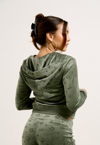 Velvet hoodie in olive green with a textured pattern featuring crowns, cherries, and symbols. Includes a hood and front pockets.
